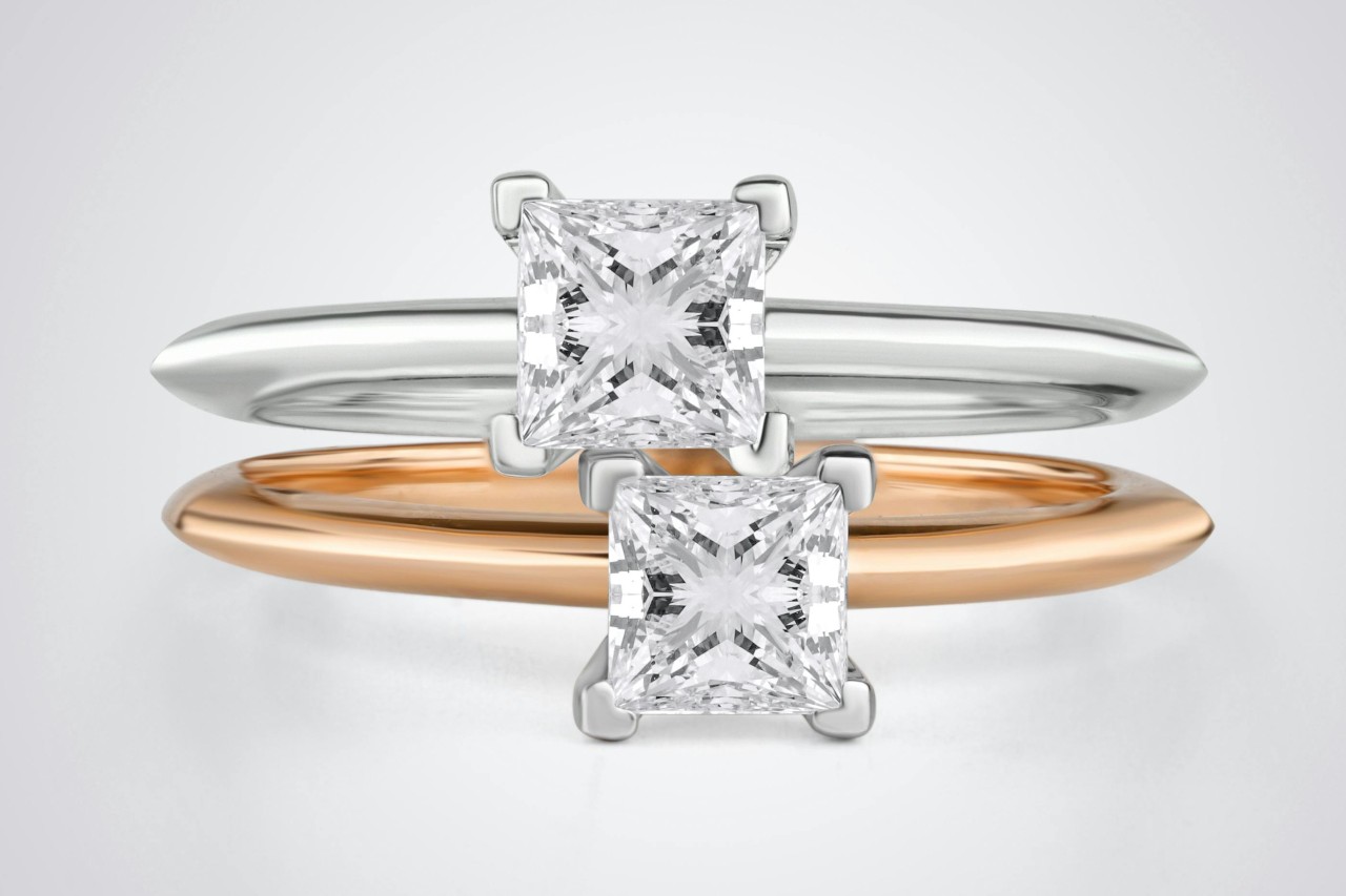 Two solitaire, princess cut engagement rings, one in white gold, the other in rose gold, stacked one on top of the other. Two solitaire, princess cut engagement rings, one in white gold, the other in rose gold, stacked one on top of the other.
