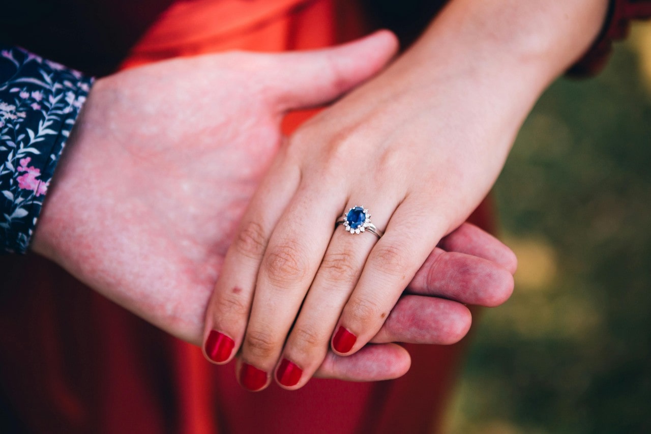 A man’s hand holding a woman’s hand, adorned with a sapphire, halo set engagement ring. A man’s hand holding a woman’s hand, adorned with a sapphire, halo set engagement ring.