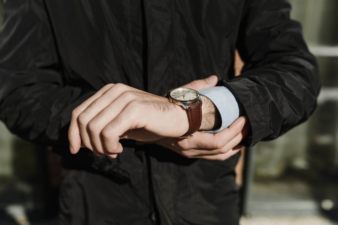 A person wearing a dark jacket checks their wristwatch, which has a brown leather strap and a white face. A person wearing a dark jacket checks their wristwatch, which has a brown leather strap and a white face.