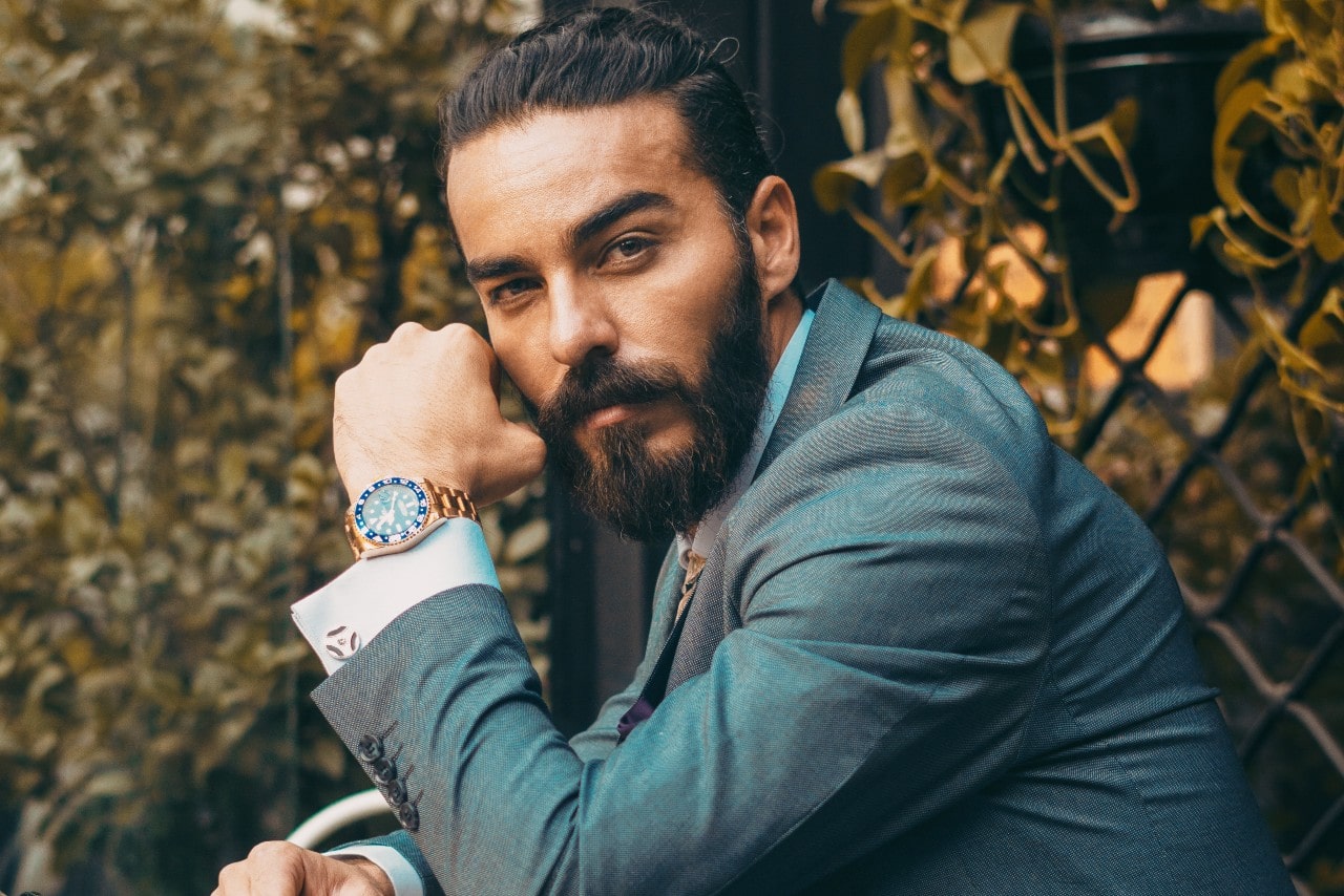 A bearded man in a blue suit gazes thoughtfully at the camera, showcasing a luxury watch. He's seated outdoors, with lush foliage in the background. A bearded man in a blue suit gazes thoughtfully at the camera, showcasing a luxury watch. He's seated outdoors, with lush foliage in the background.