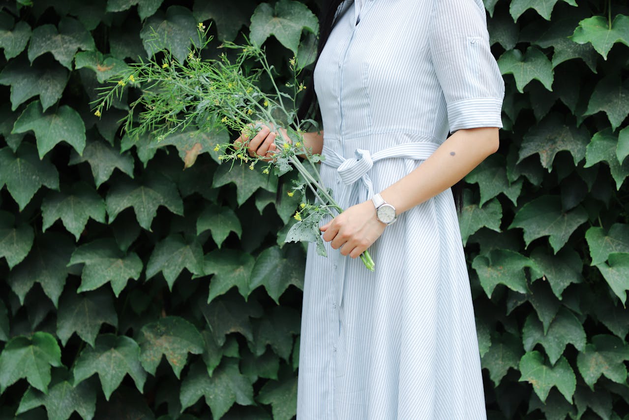 A close-up of a woman standing in front of a wall of ivy, wearing a simple watch and holding a sprig of small yellow flowers.