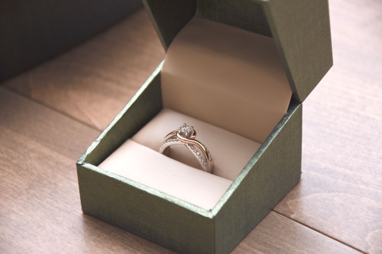 A two-tone split shank engagement ring in a ring box on a wooden table. A two-tone split shank engagement ring in a ring box on a wooden table.